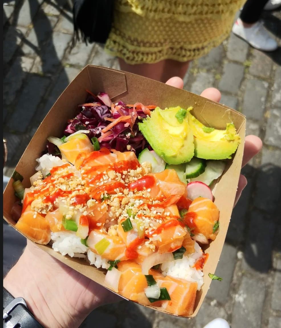 Salmon poke bowl topped with spicy sauce, avocado, and red cabbage in a cardboard container.