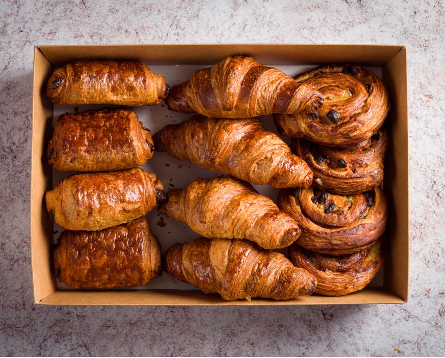 Cardboard box filled with golden brown croissants, pain au chocolat, and swirled raisin pastries.