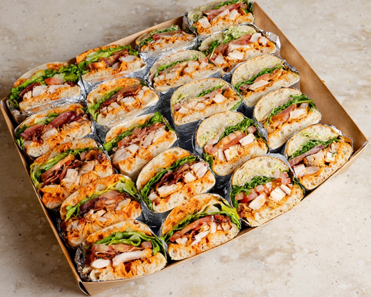 Cardboard catering box filled with chicken, bacon, and lettuce sandwich halves wrapped in foil.