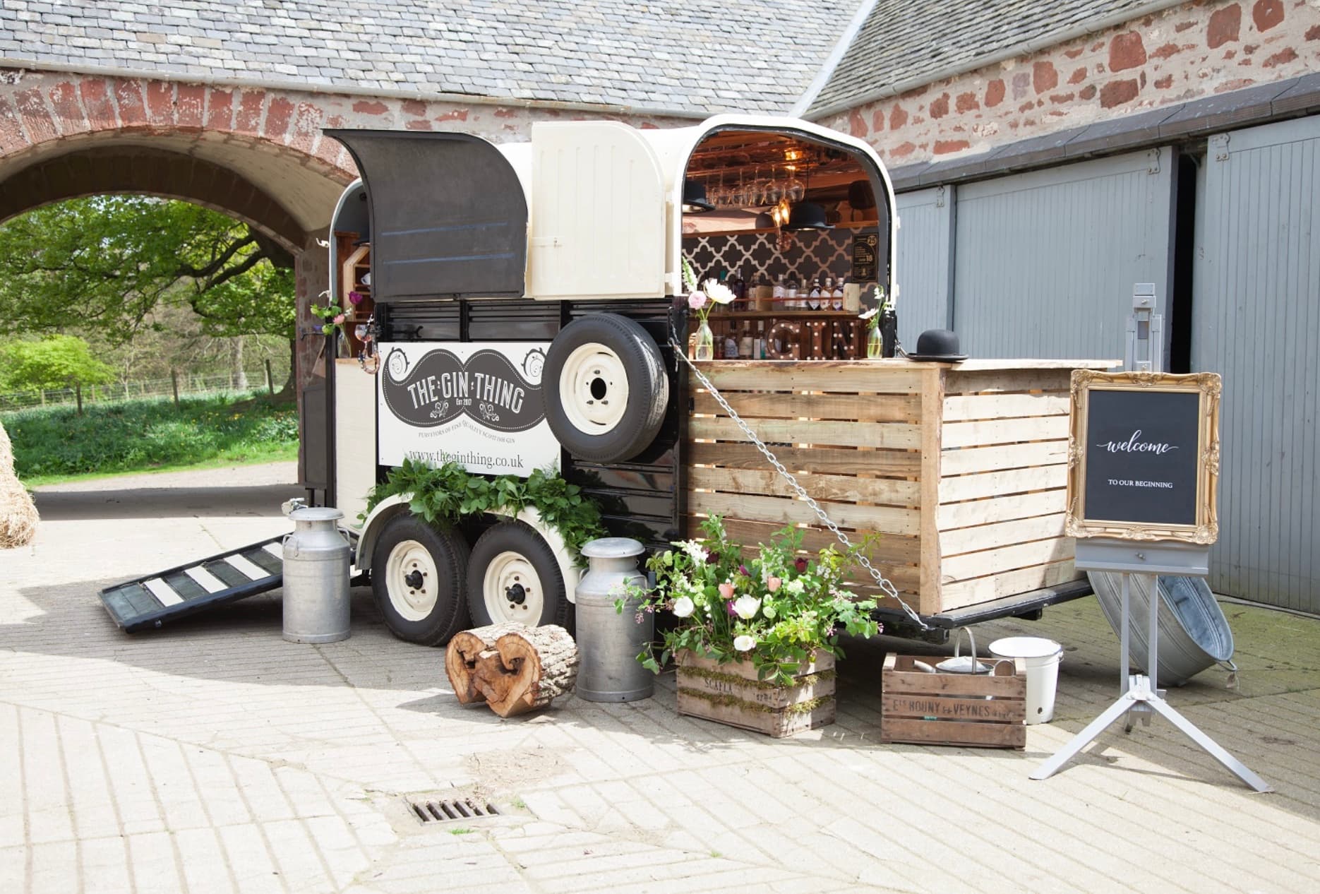 Rustic mobile gin bar converted from a horse trailer, featuring wooden counters and floral arrangements.