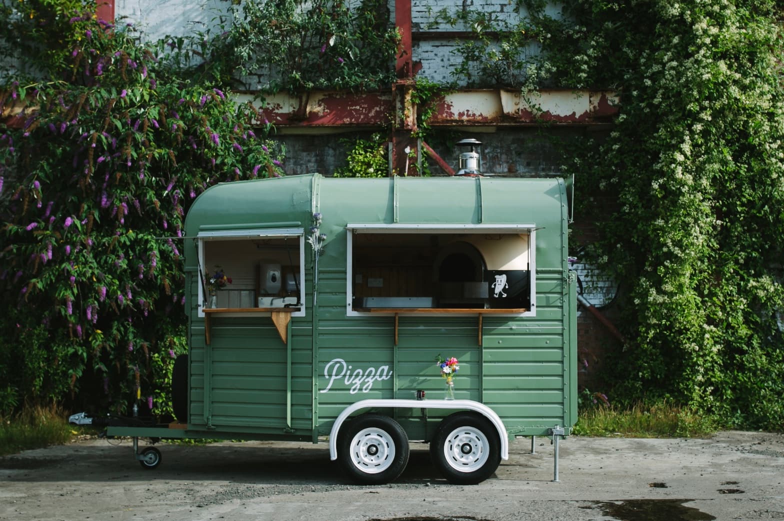 Sage green horse trailer converted into a mobile pizza kitchen parked beside lush greenery.