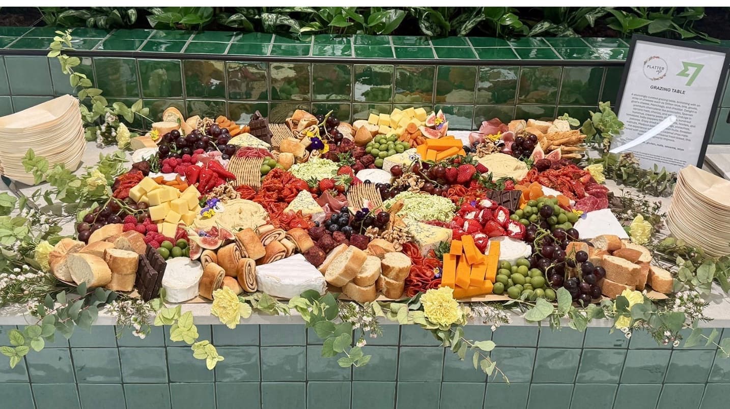 Colorful grazing table with assorted cheeses, meats, fruits, and bread on a green tiled counter.