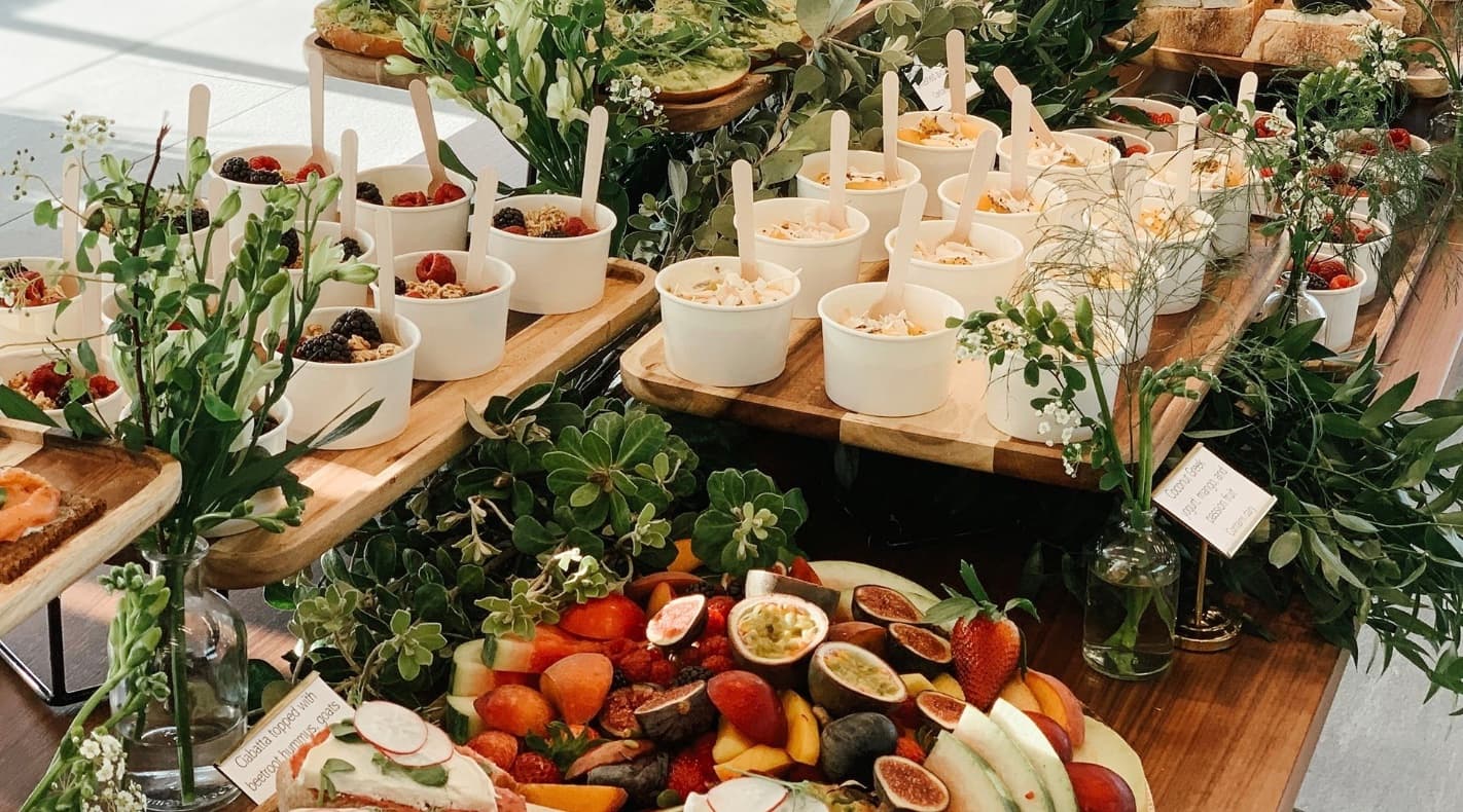 Breakfast buffet spread with yogurt cups, fresh fruit platters, and decorative greenery on wooden boards.